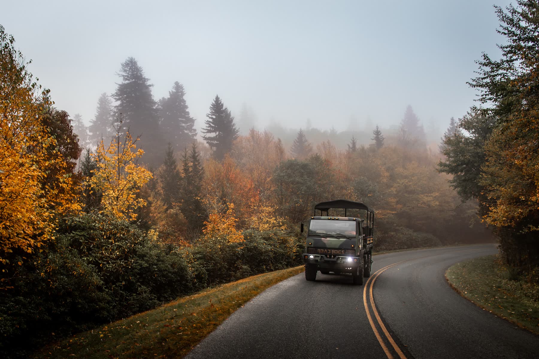 The restored military transport driving a fog-covered mountain road in peak autumn colour, headlights on