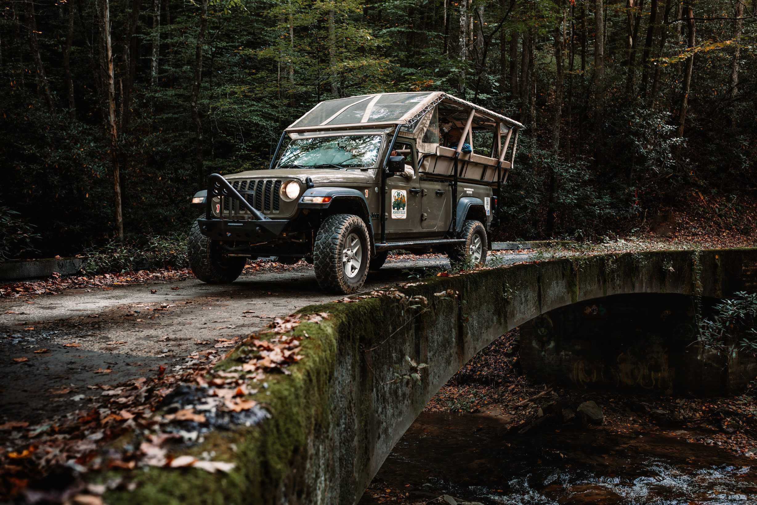 Open-air Jeep crossing a moss-covered stone bridge over a running creek on a forest backroad