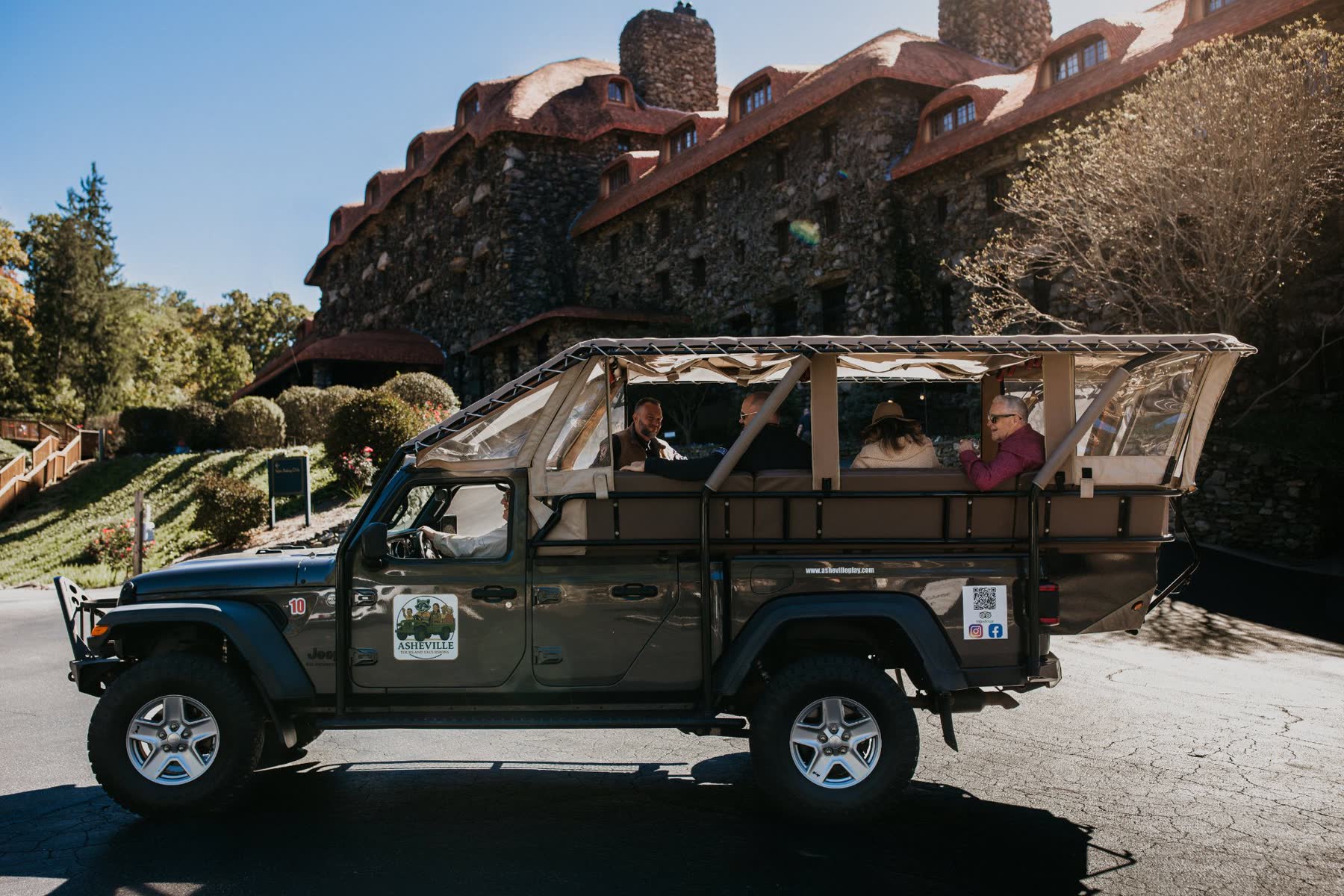 Guests seated in the elevated open-air bed of the safari Jeep outside a historic stone lodge